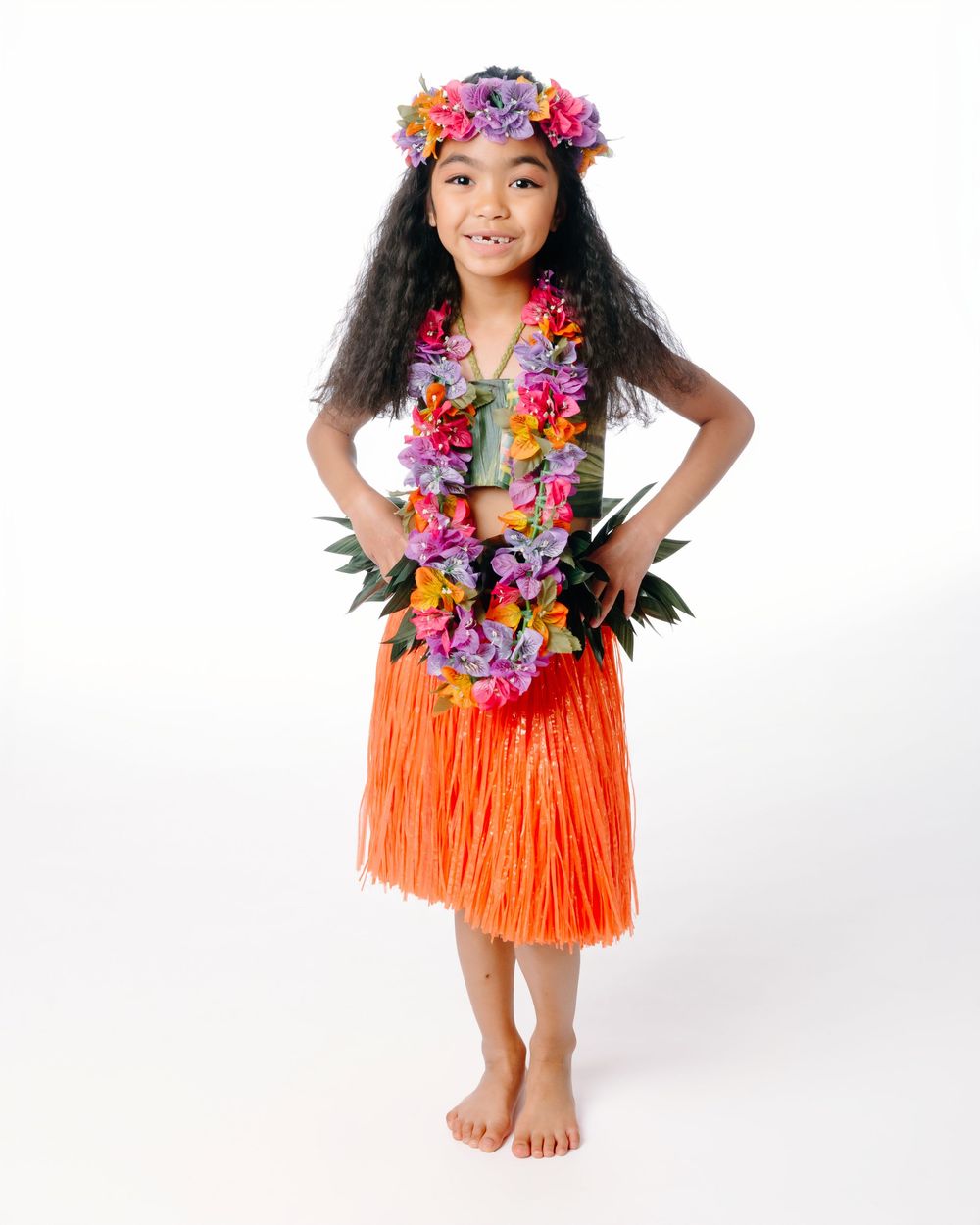 Young girl in Hawaiian hula costume with orange grass skirt, colorful lei, and flower crown on white background.