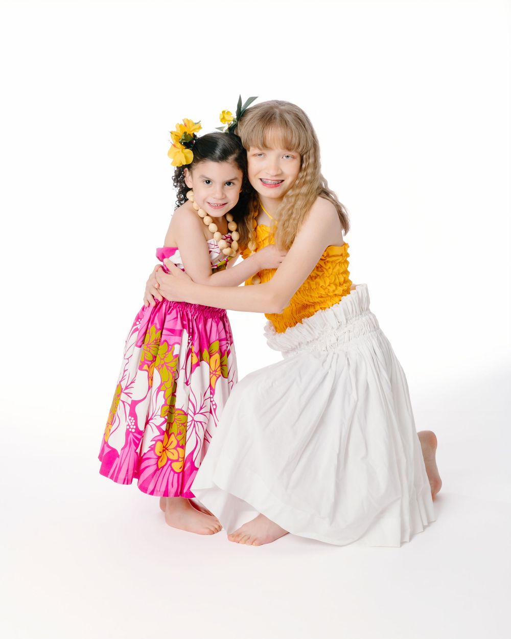 Two young girls in colorful Hawaiian hula outfits hugging and smiling against a white background.
