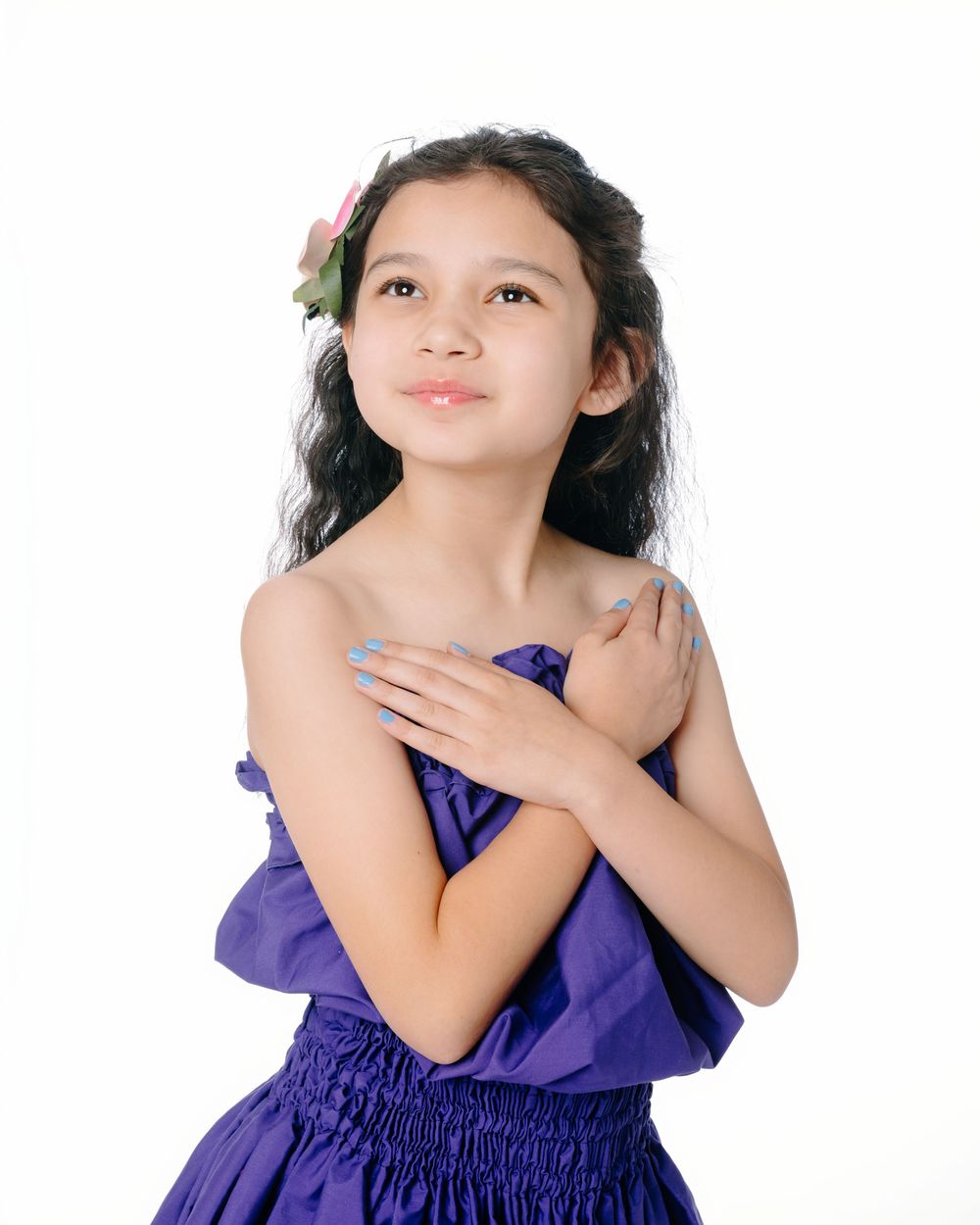 Young girl in purple dress with flower hair accessory, hands crossed over chest, looking upward on white background.
