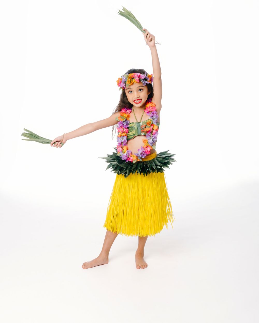 Young girl in yellow hula skirt, floral lei, and headpiece dancing with green leaves on white background.