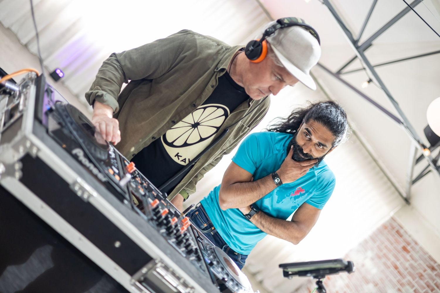A man plays music on a DJ mixing desk whilst another man looks on