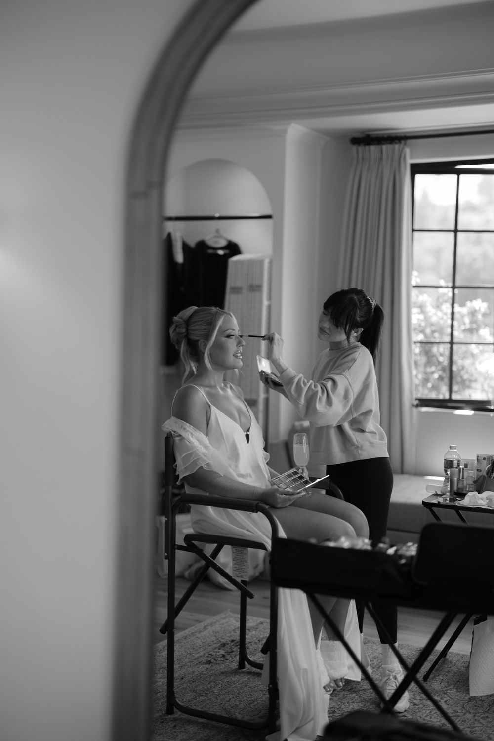 Black and white photo of a makeup artist applying cosmetics to a client seated in front of a window in an elegant room.