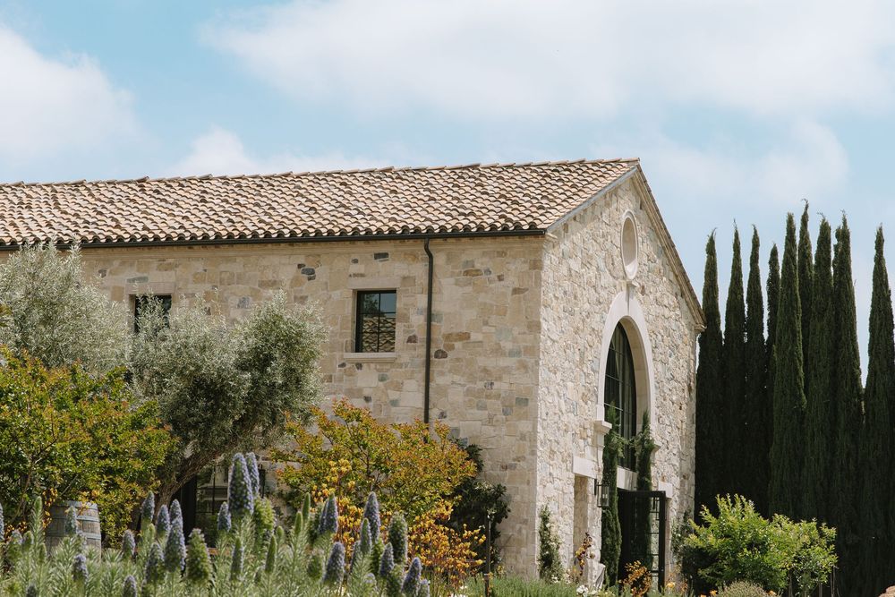 Mediterranean stone building with arched doorway and cypress trees in rustic countryside setting.