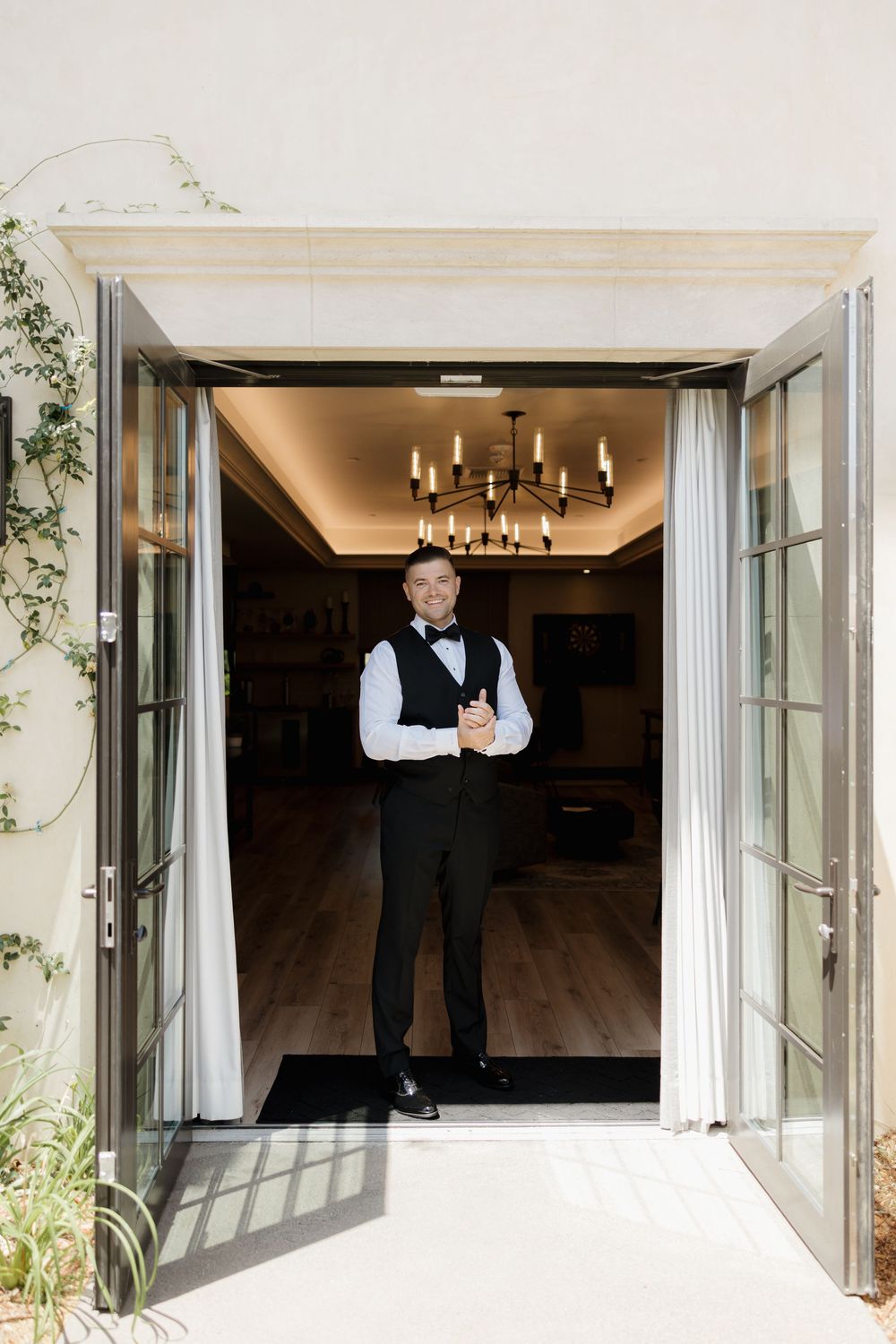 Groom in formal attire standing in doorway with elegant chandelier overhead.