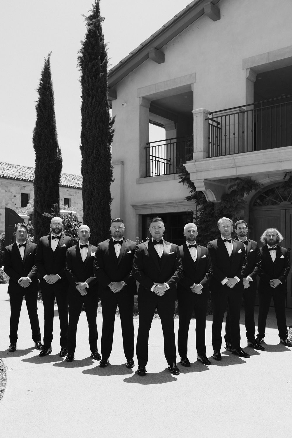Groomsmen in classic black tuxedos and bow ties lined up against a Mediterranean-style building exterior.