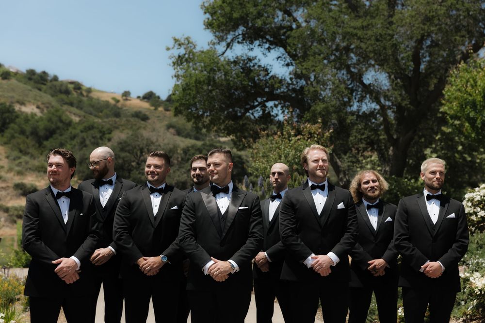 Groomsmen in classic black tuxedos and bow ties stand together outdoors against a scenic hillside backdrop.