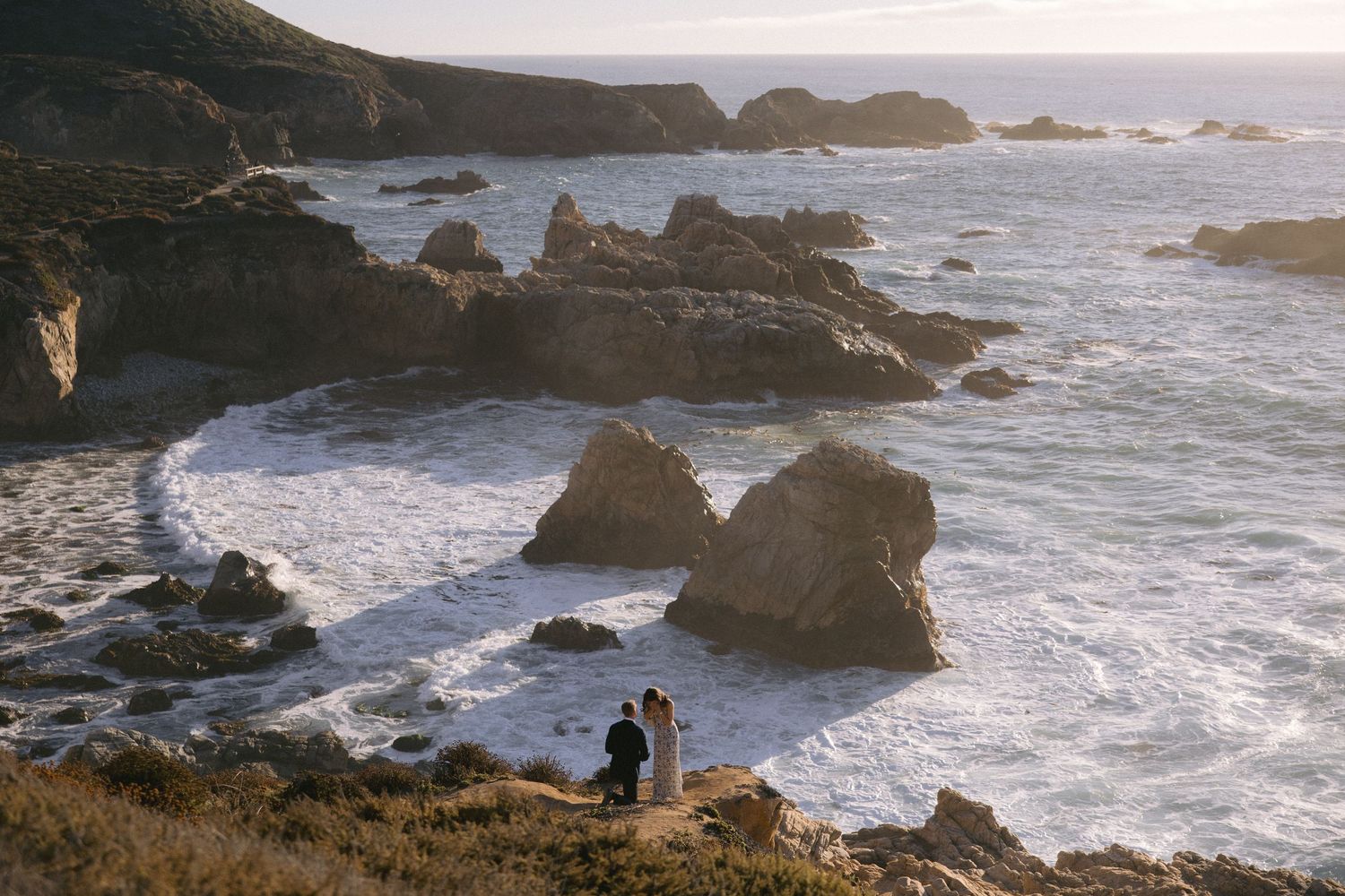 Dramatic coastal landscape with rocky cliffs and crashing waves along a rugged shoreline at sunset.
