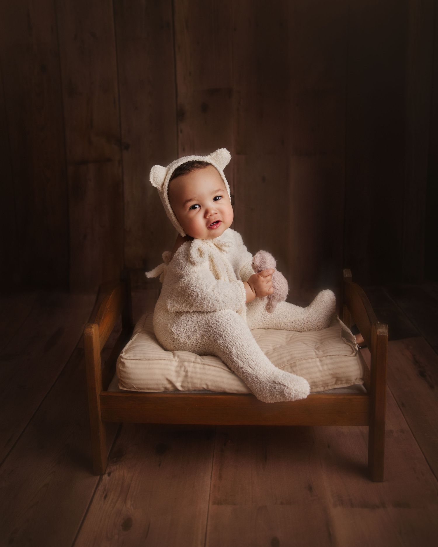 Baby sitting on little bed with teddy bear outfit holding teddy stuffy in hands