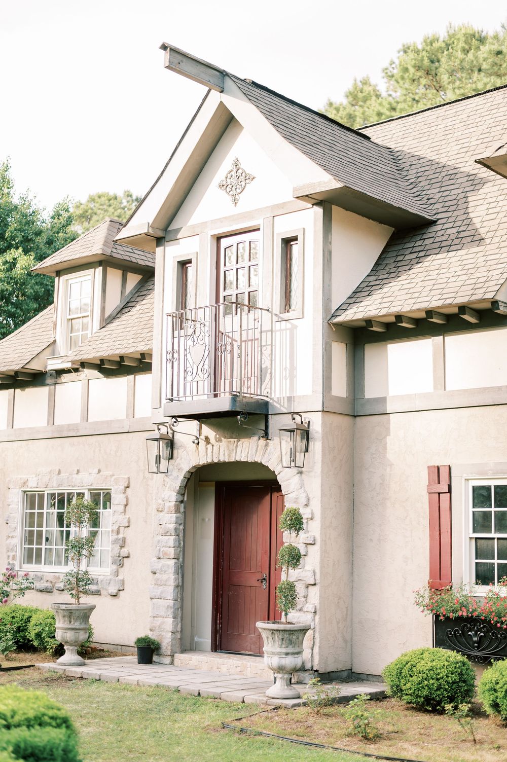 Elegant stone and stucco Tudor-style home exterior with burgundy door, ornate balcony, and manicured landscaping.