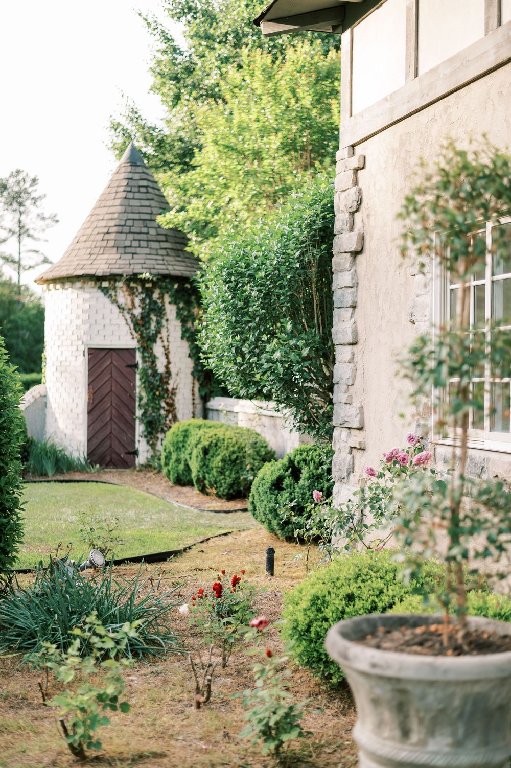A charming stone turret with brown wooden door leads to a garden path lined with lush greenery and potted plants.