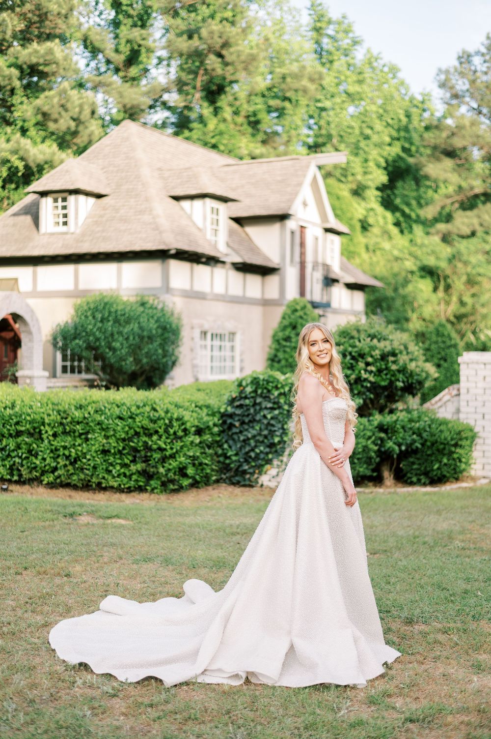 An elegant white wedding gown with a long train flows across a manicured lawn in front of a grand Tudor-style mansion.