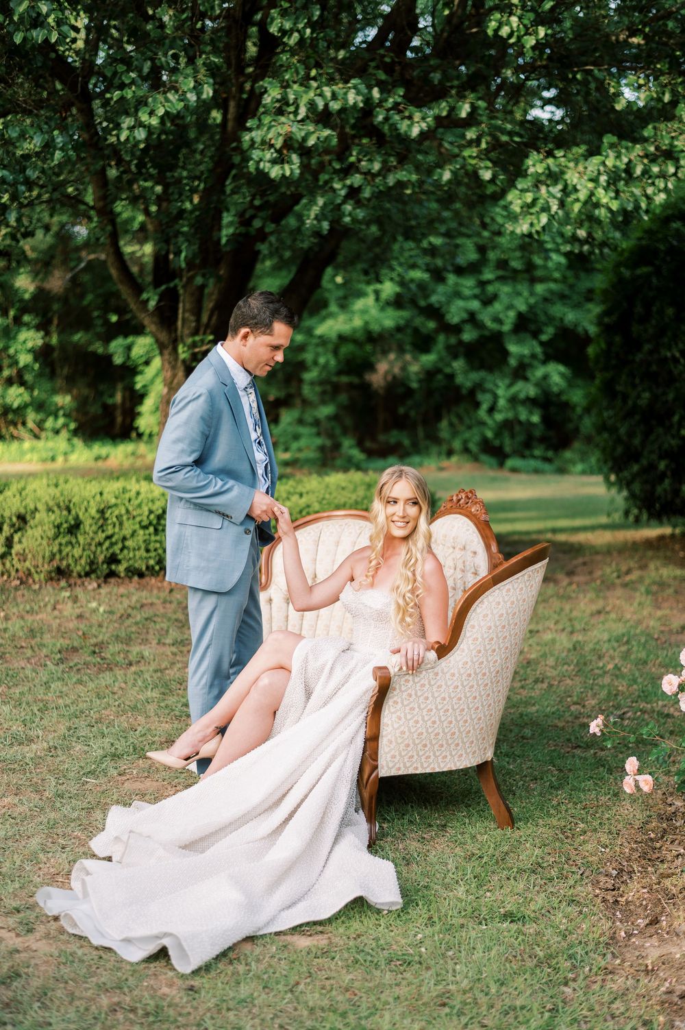 Couple poses outdoors in wedding attire with bride seated in vintage chair on garden lawn surrounded by greenery.
