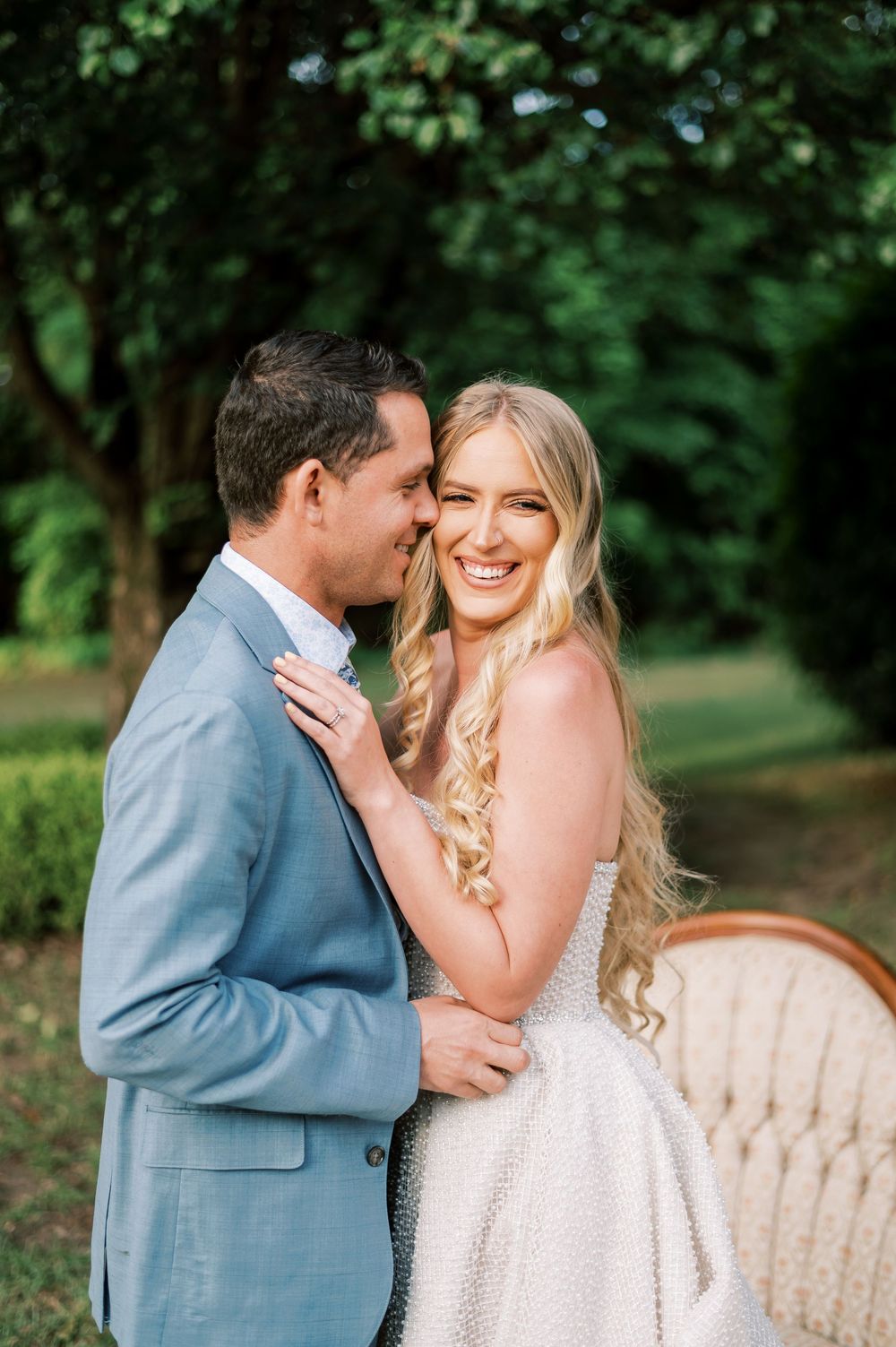 A couple embraces outdoors in a garden setting with a vintage couch, bride in white dress and groom in light blue suit.