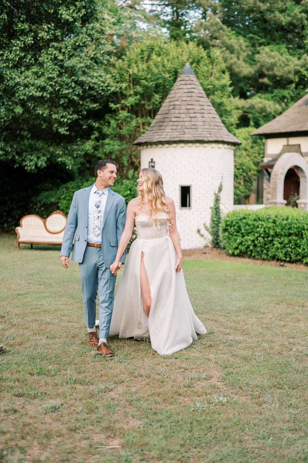A couple walking hand in hand across a garden lawn with a charming stone turret and landscaping in the background.