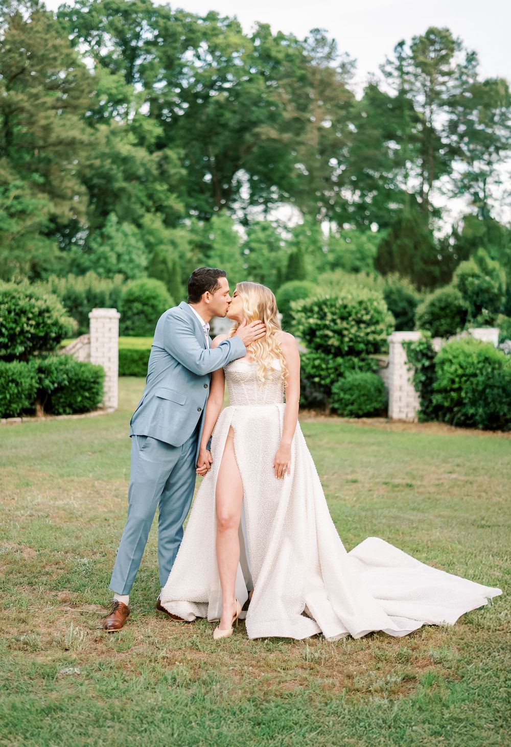 A romantic wedding photo captured in a lush garden setting with stone pillars and flowing white gown.