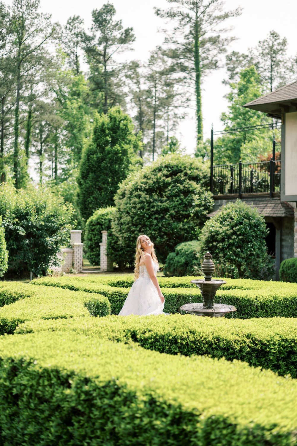 Figure in a formal gown walks through manicured garden hedges surrounded by tall pine trees and ornate landscaping.