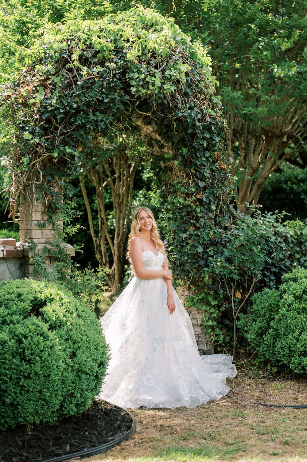 A person in an elegant white lace wedding gown poses under a lush garden archway covered in green vines and foliage.