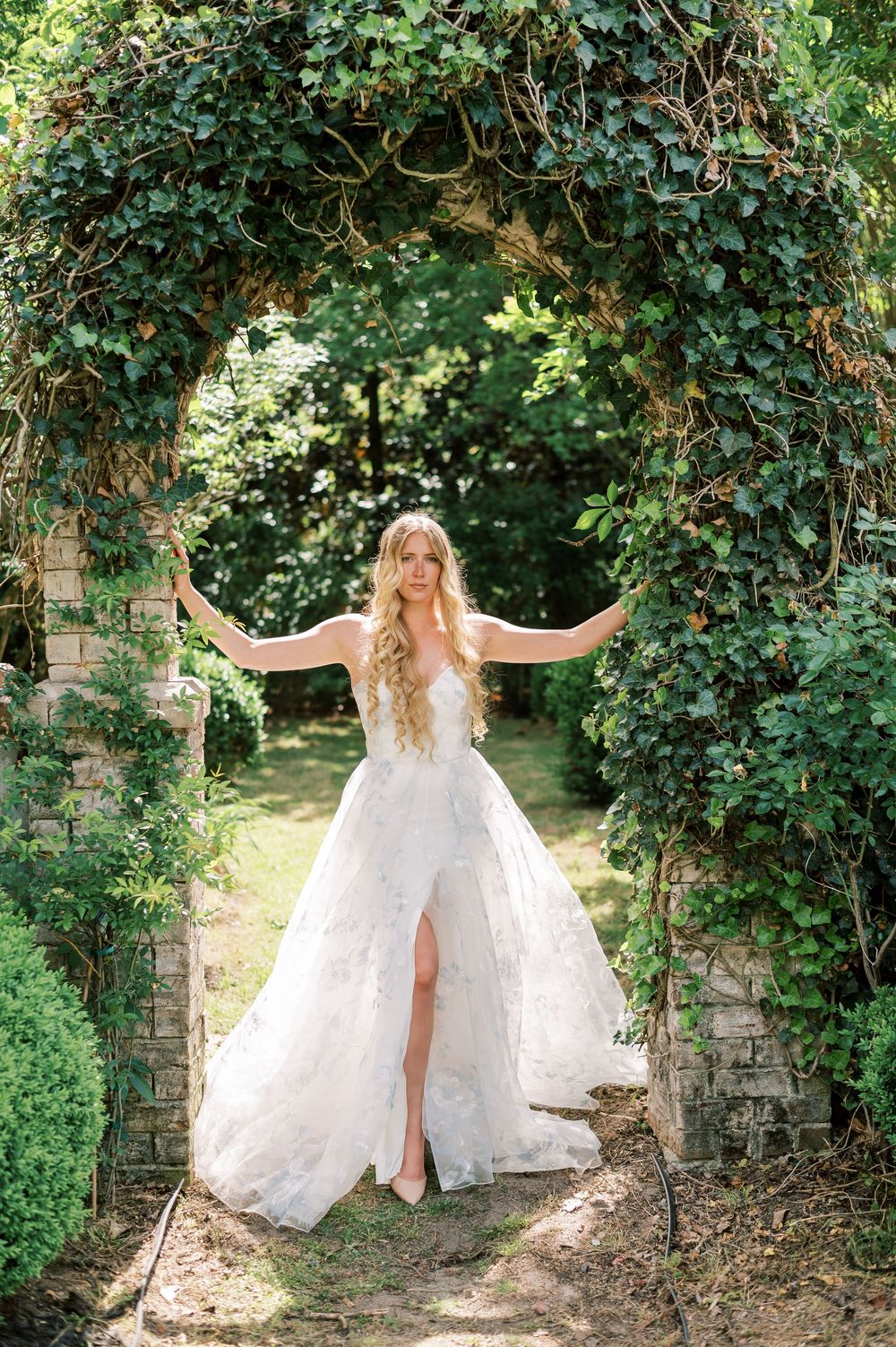 A romantic garden archway covered in lush green ivy frames a white wedding gown against a sunlit natural backdrop.