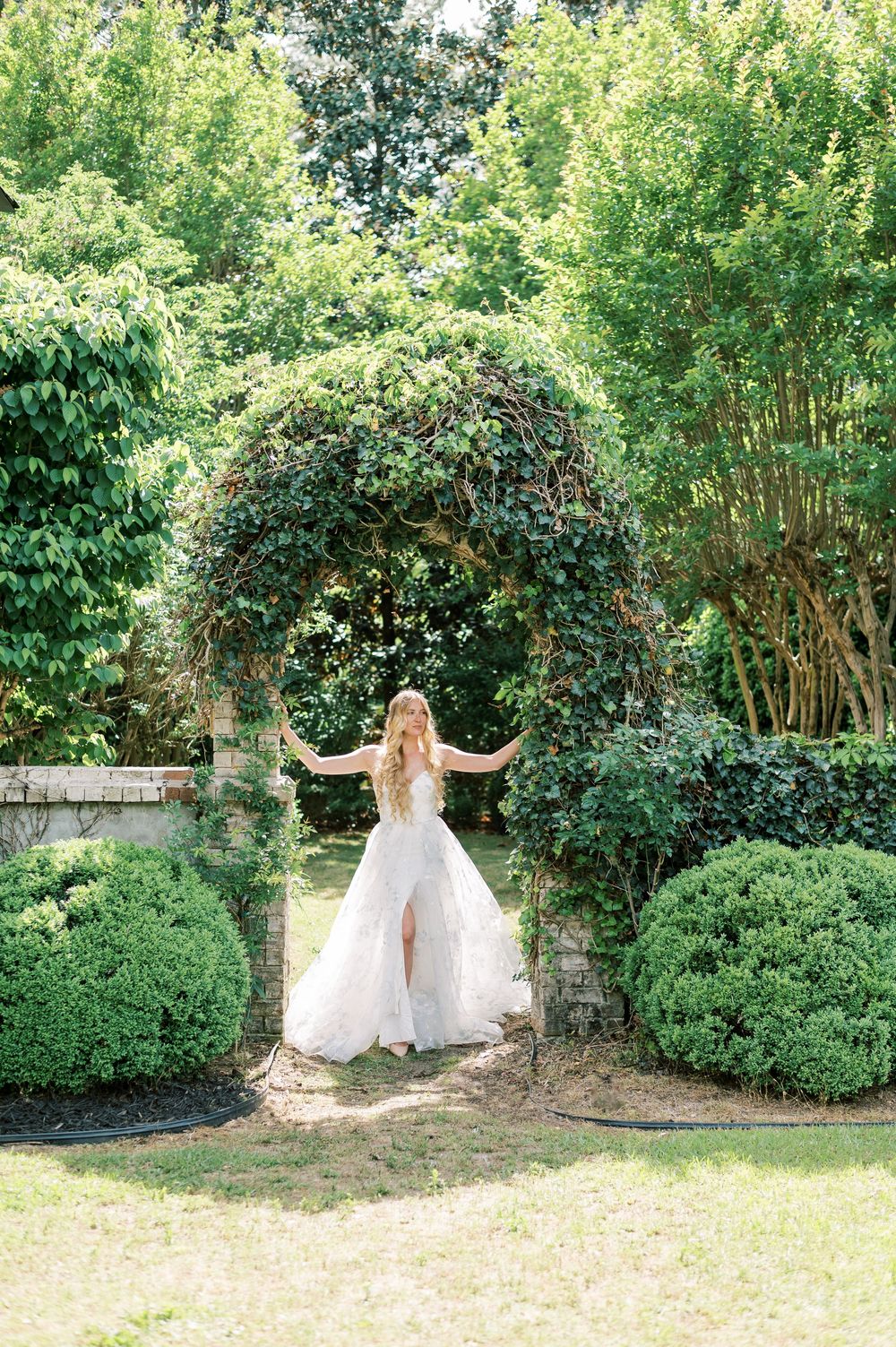 A flowing white wedding gown captured in a lush garden setting with an ivy-covered archway and manicured boxwood hedges.