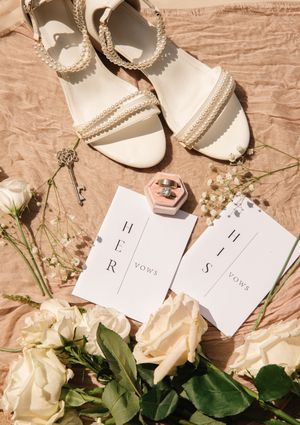 Wedding details flatlay with white sandals, vow books, roses and delicate accessories on blush fabric.