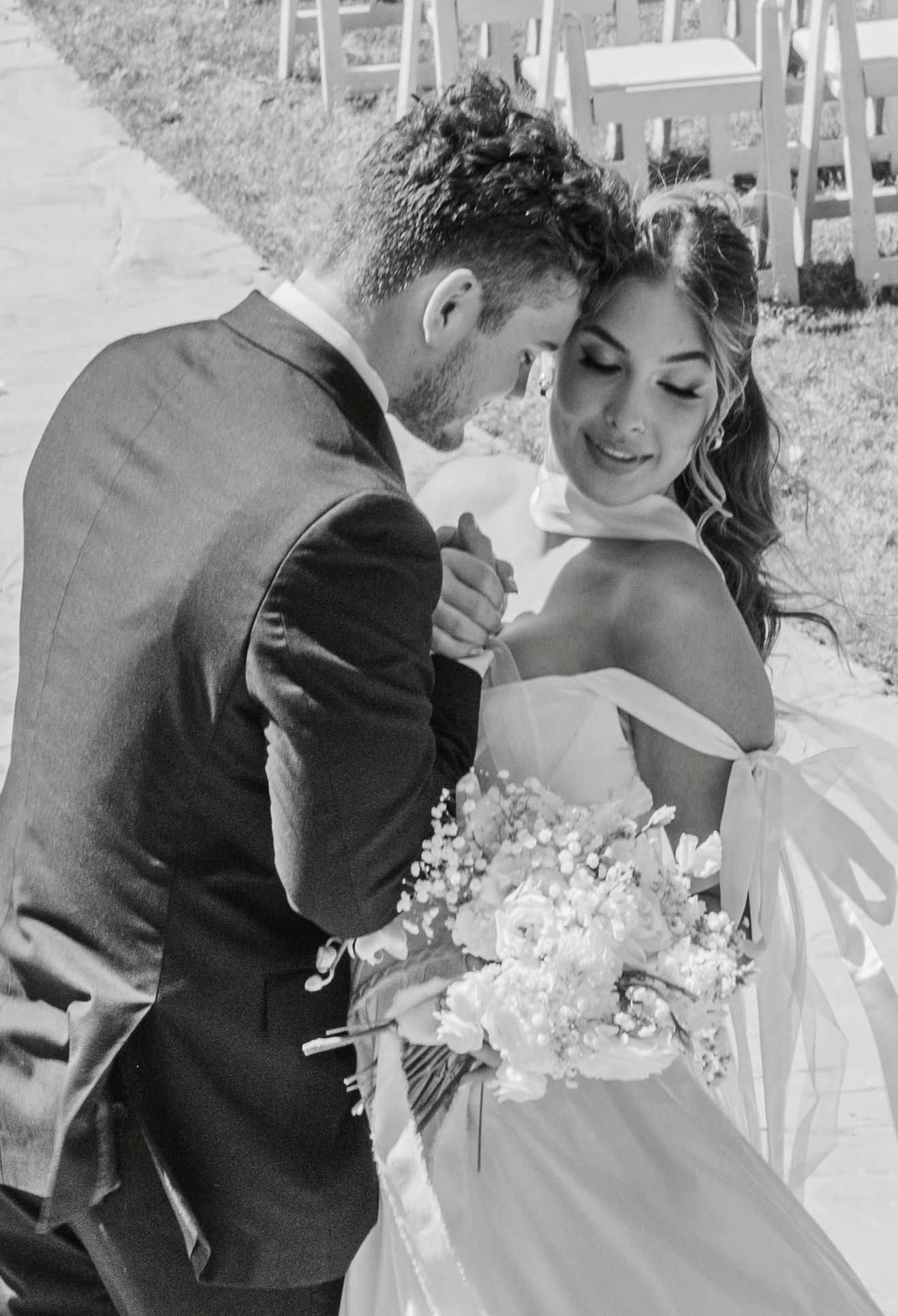 A romantic black and white wedding moment captured outdoors with white chairs and a delicate bouquet in view.