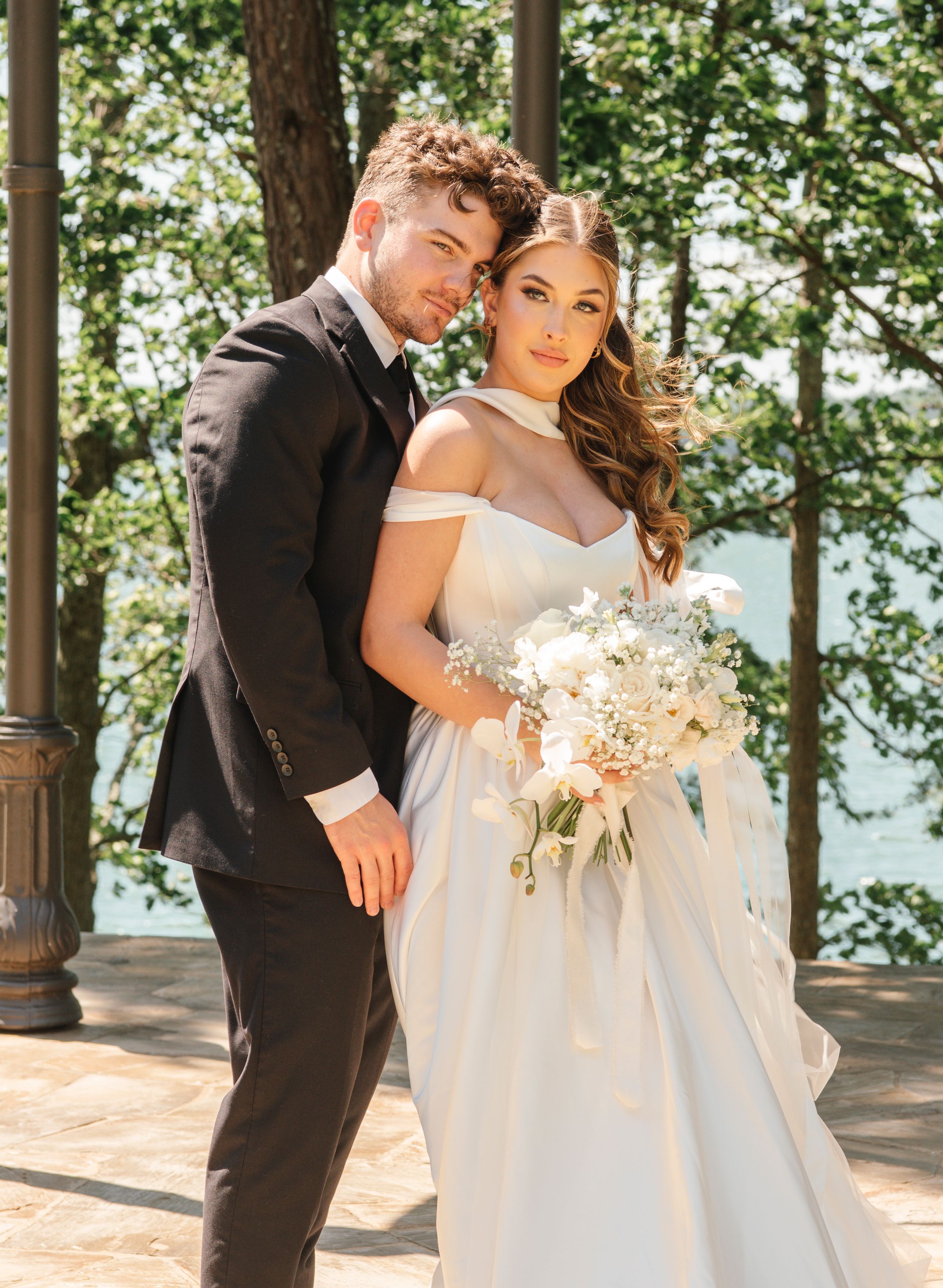 A romantic wedding portrait of a couple in formal attire standing outdoors with a white floral bouquet.