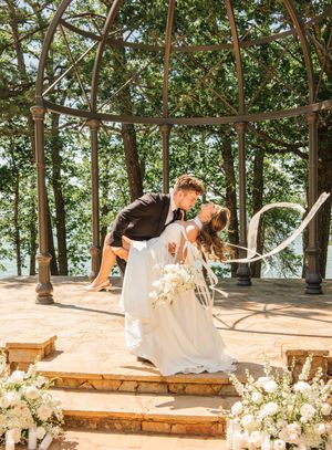 A romantic wedding moment under a gazebo with white floral decorations and wooden steps surrounded by lush trees.
