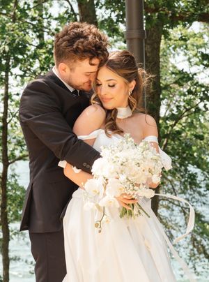 Romantic wedding portrait of newlyweds embracing outdoors with bride holding white floral bouquet in lace gown.