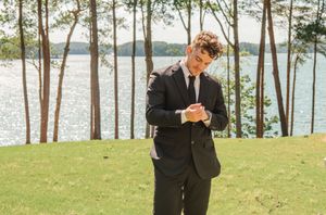 Groom in black suit checks time while standing near lakeside with pine trees.