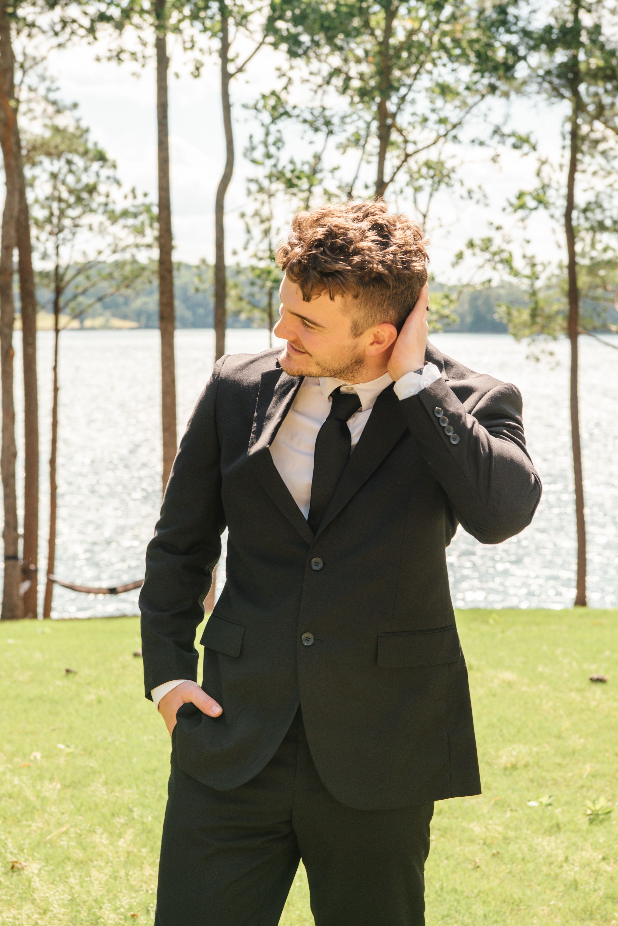 A well-dressed person in a black suit stands by a lakeside on a grassy lawn among trees.