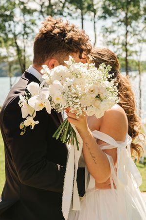 A couple embraces at their outdoor wedding as the bride holds a white orchid and baby's breath bouquet by a lakeside.