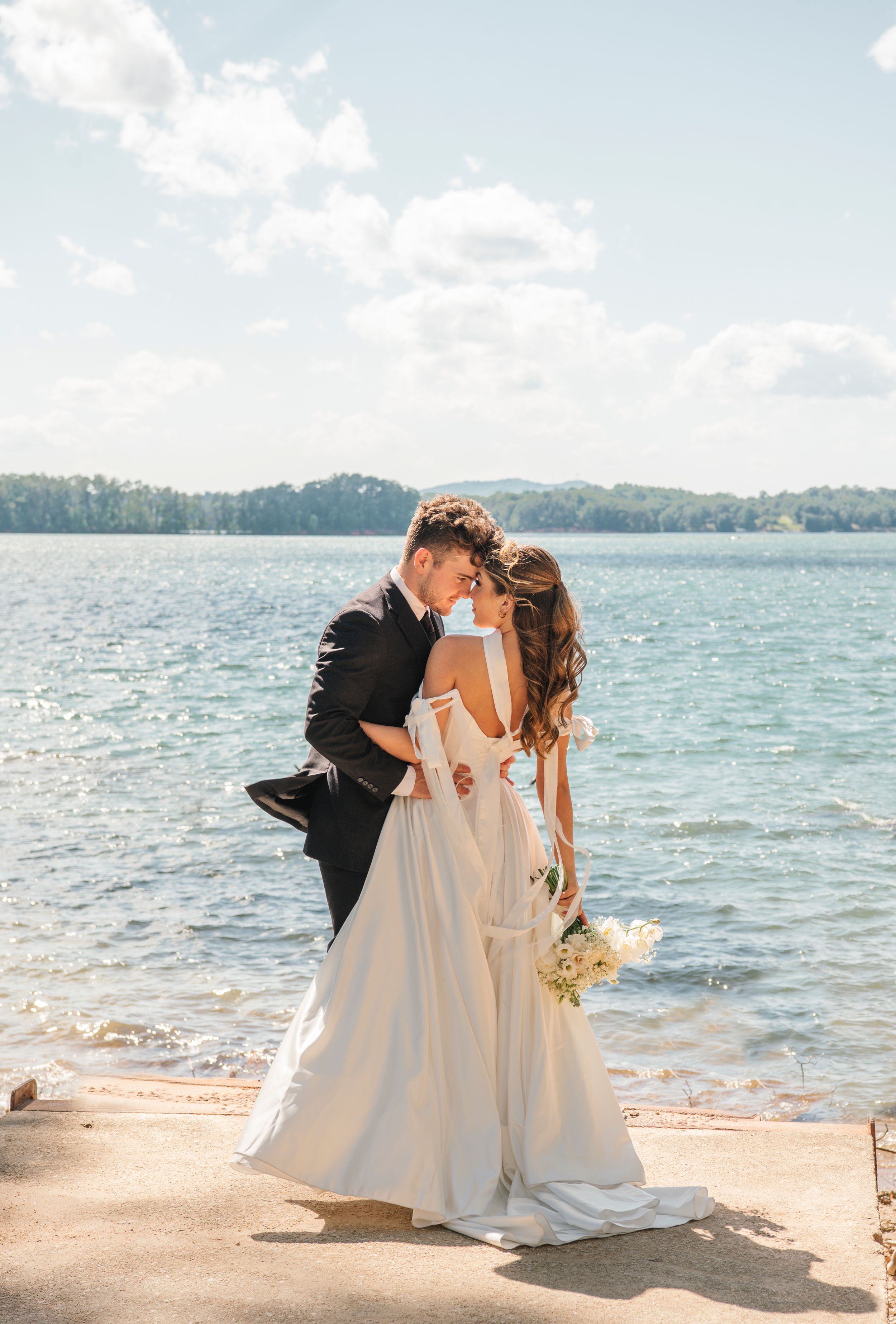Newlyweds share intimate moment by lakeside on sunny wedding day with white dress and black suit.