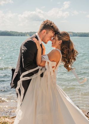 Newlywed couple embraces tenderly on a beach shoreline with sparkling water and cloudy sky in the background.