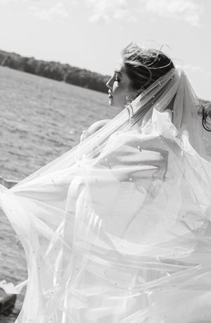 Black and white portrait of bride with flowing veil against lake backdrop in dramatic pose.