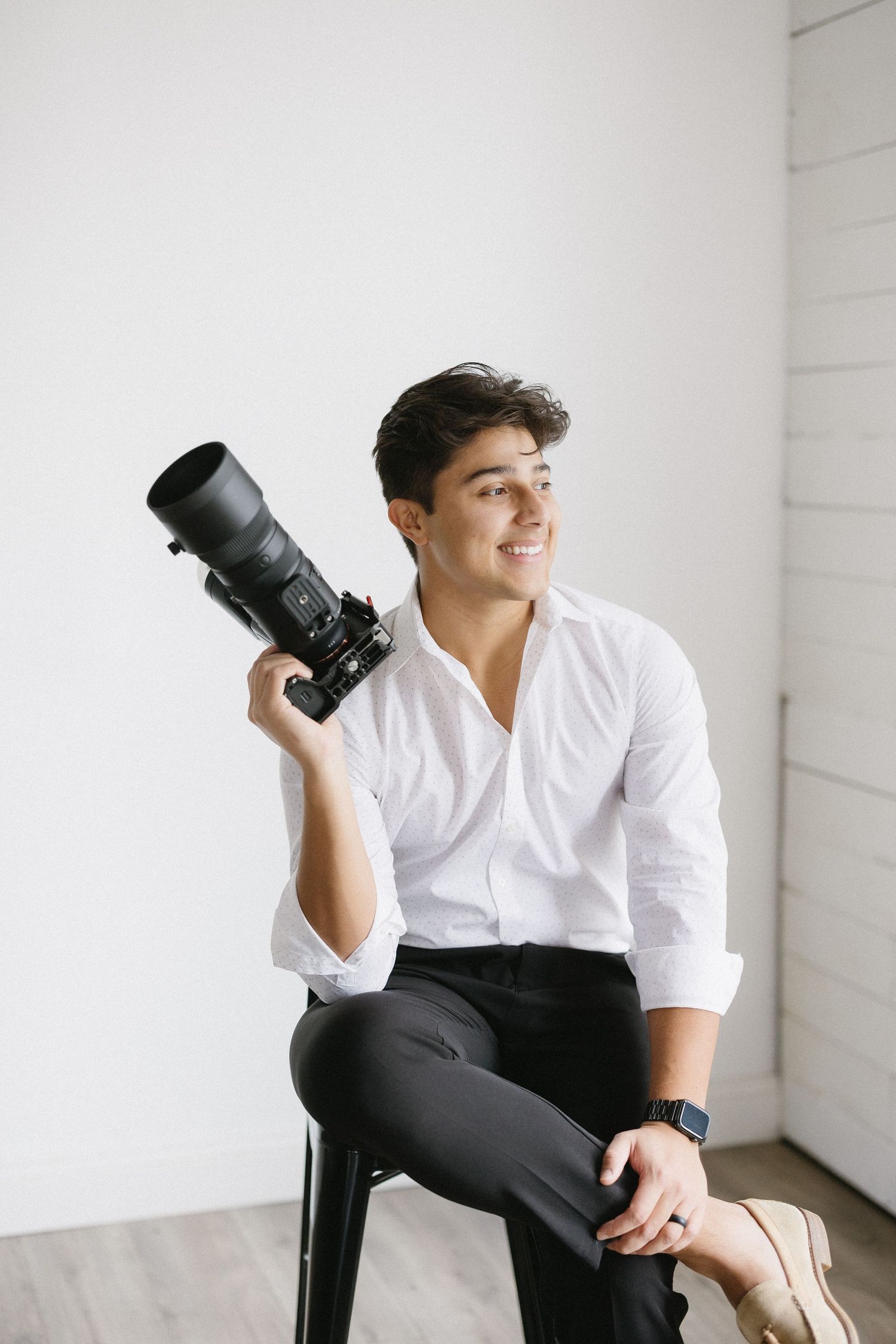 Professional photographer in white shirt holding camera while sitting on stool.