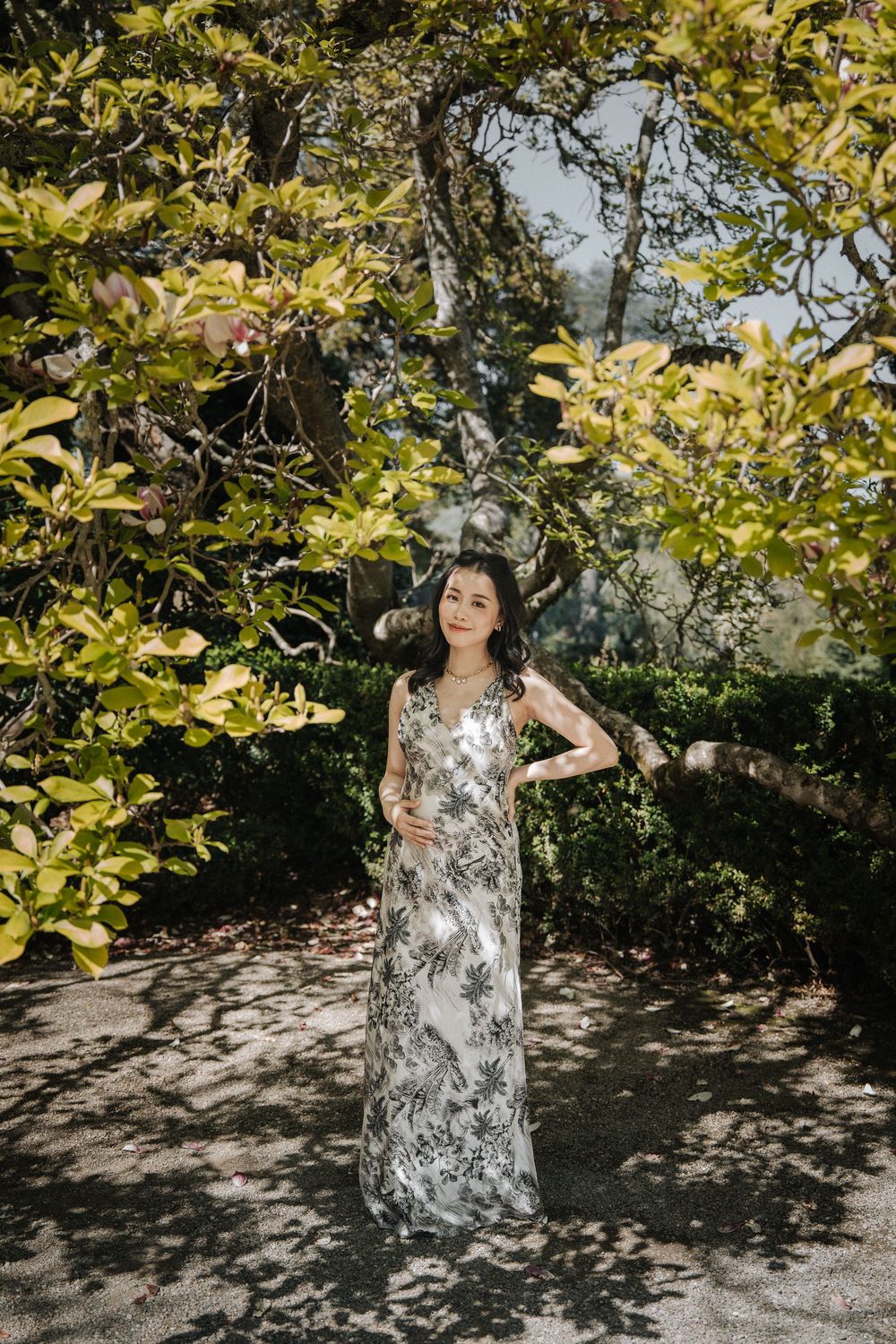 Person in floral white dress poses gracefully under tree branches with dappled sunlight.
