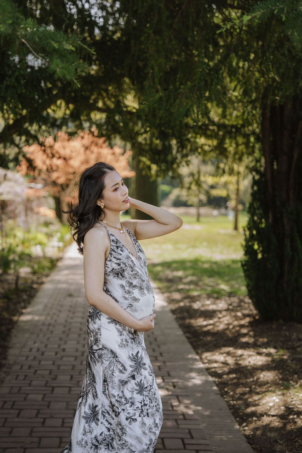 Person in white floral dress poses playfully on brick pathway surrounded by trees.