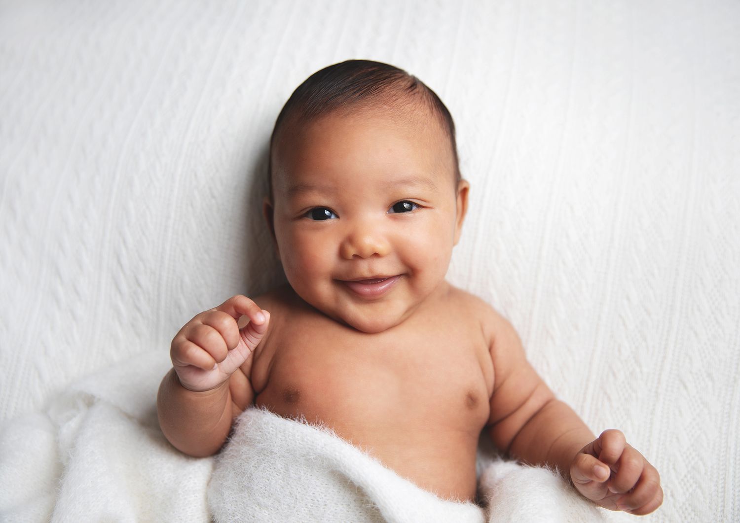 Smiling baby lying on soft white blanket showing bright expression. During a newborn photoshoot in Bexley