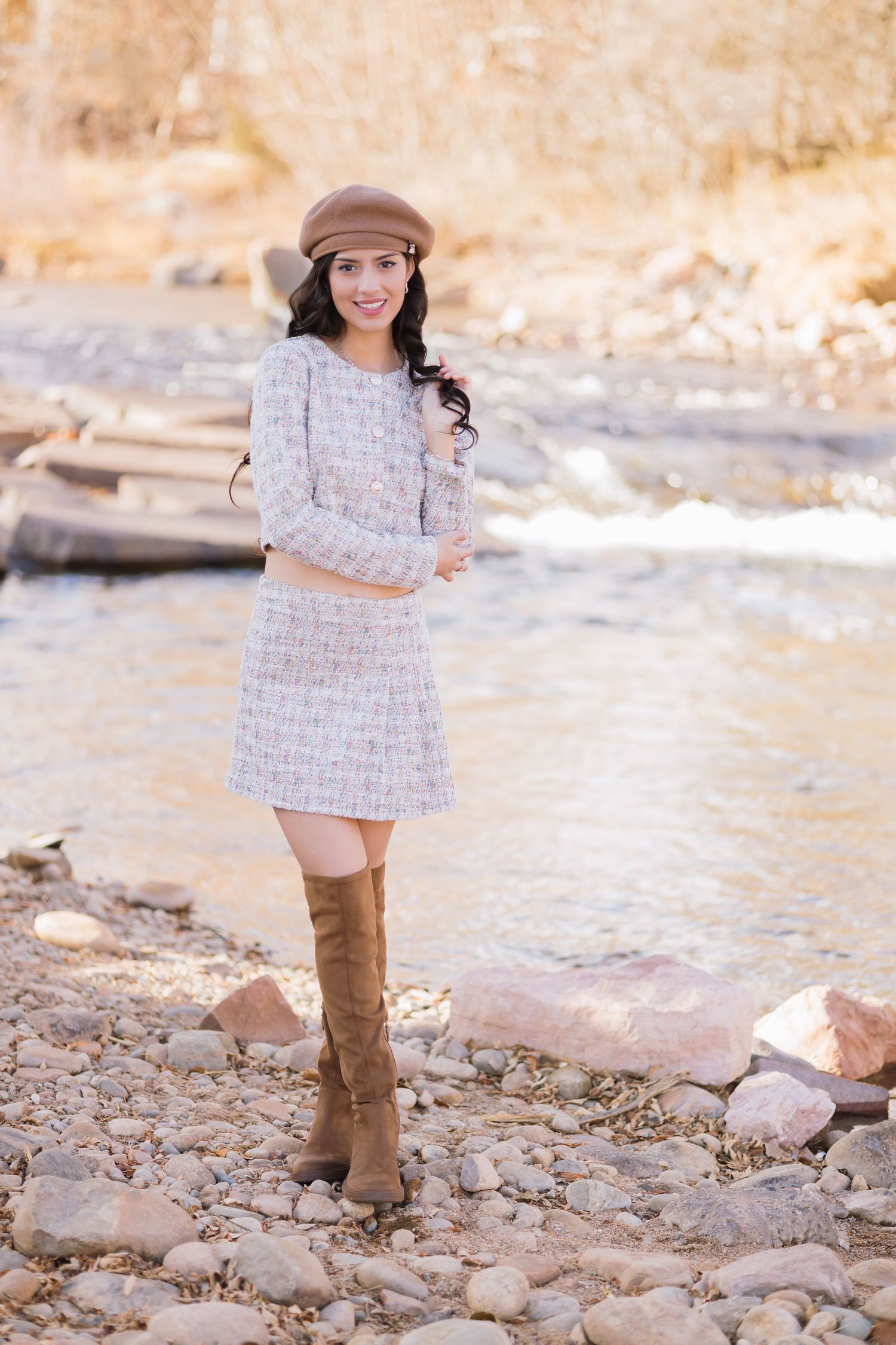 Someone in a tan newsboy cap, white tweed dress and brown knee-high boots poses on a rocky riverbank at sunset.
