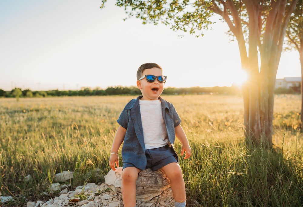 Small child in sunglasses and casual clothes sits on a rock in a sun-lit meadow at golden hour.
