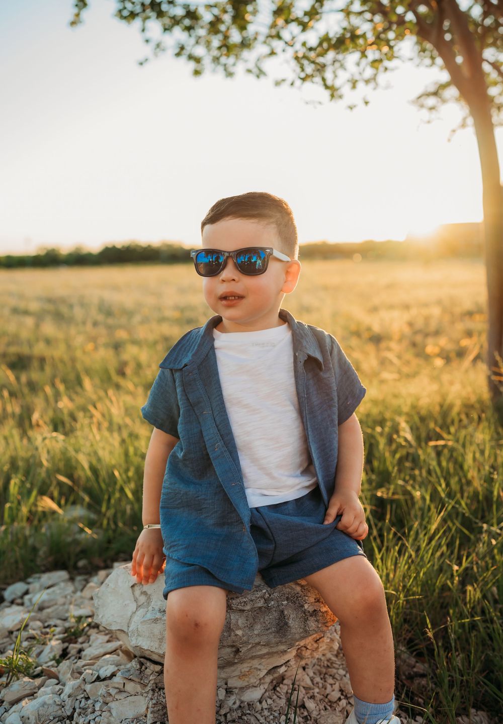 Young child wearing sunglasses and denim outfit sits on rock during golden hour sunset in grassy field.