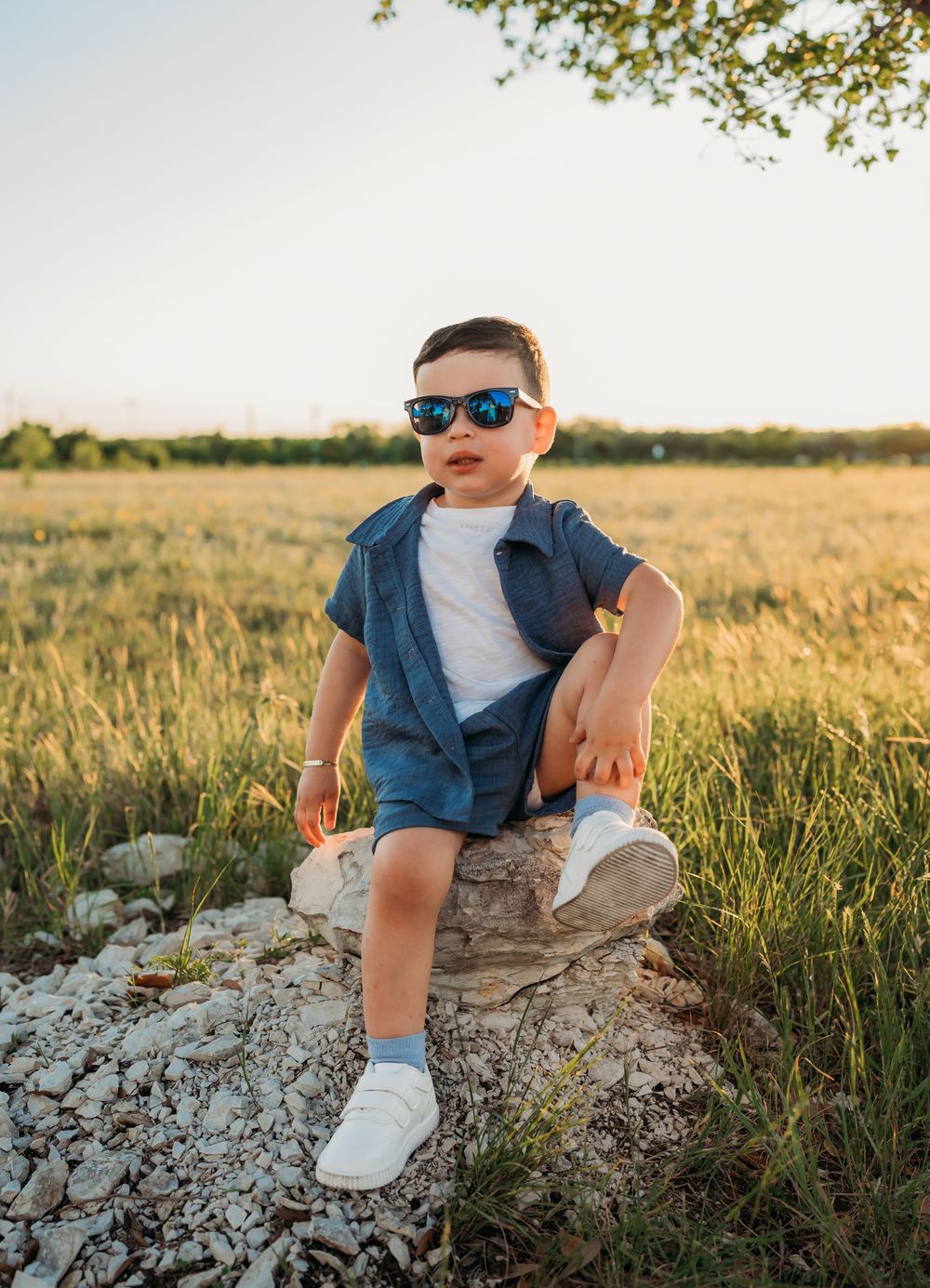 Young child in sunglasses and casual summer outfit sits confidently on a rock in a sunny meadow at golden hour.