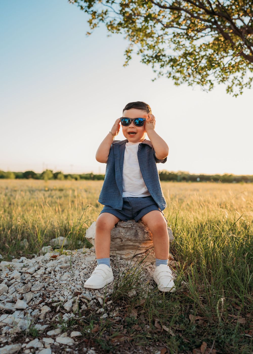 Small child wearing sunglasses and vest sits on rock in golden field at sunset with tree branches overhead.