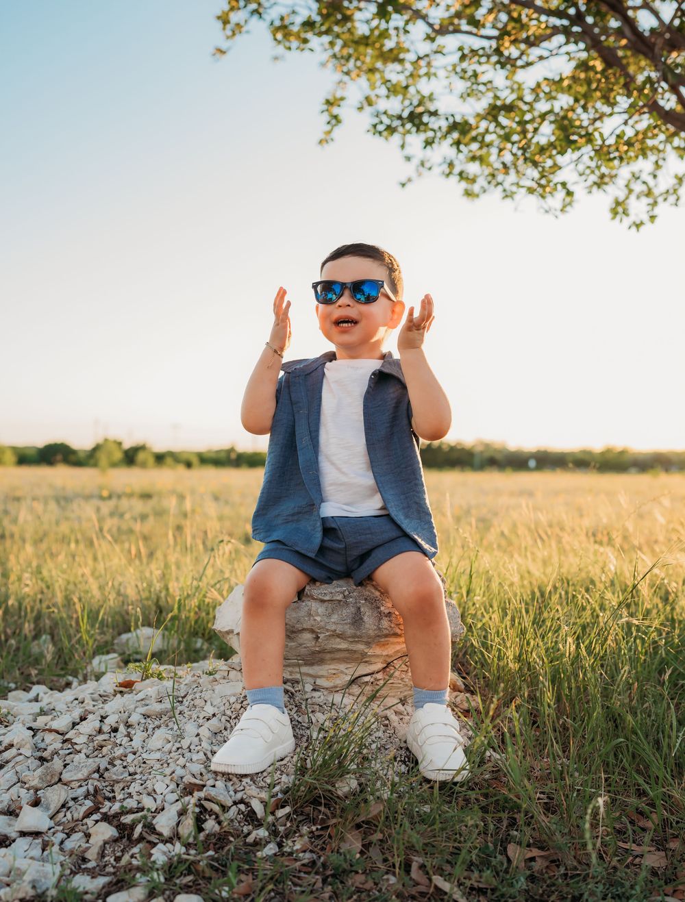 Small child in sunglasses and casual outfit sits on a rock in a sunny field with raised hands expressing joy.