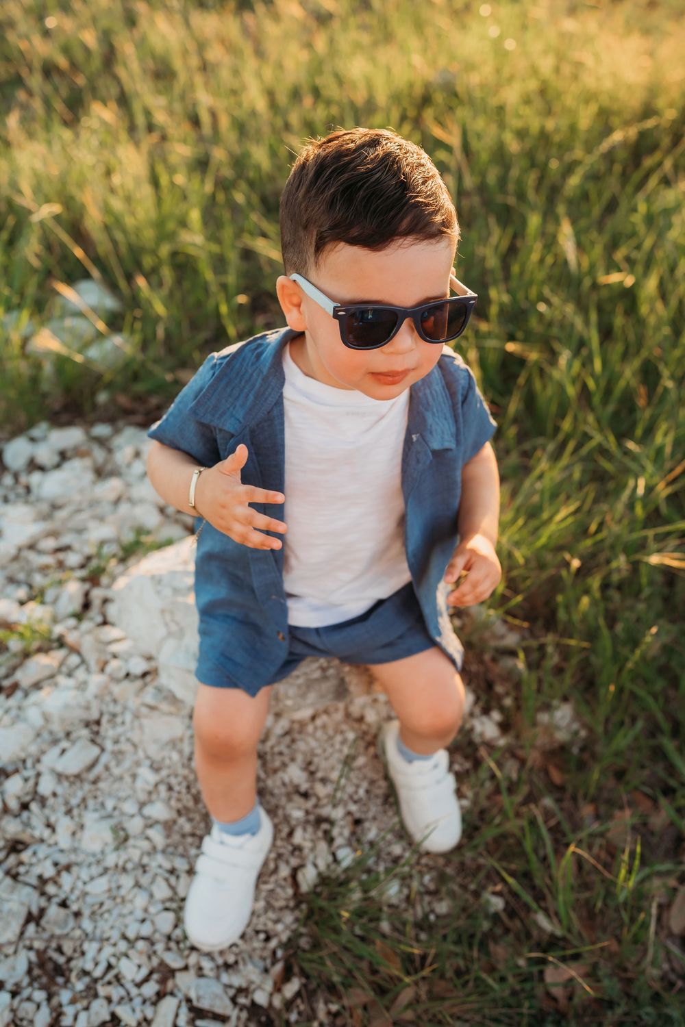 A young child in sunglasses and denim outfit strikes a cool pose while standing on rocky ground at golden hour.
