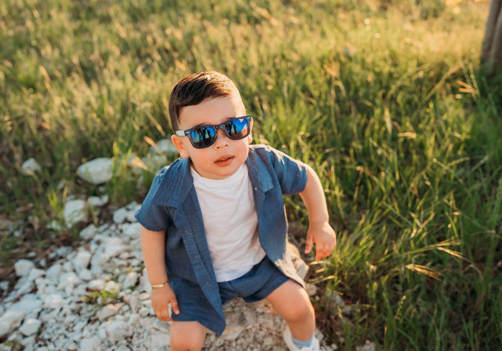 Young child wearing sunglasses, navy blazer and white shirt poses stylishly in grassy field on sunny day.