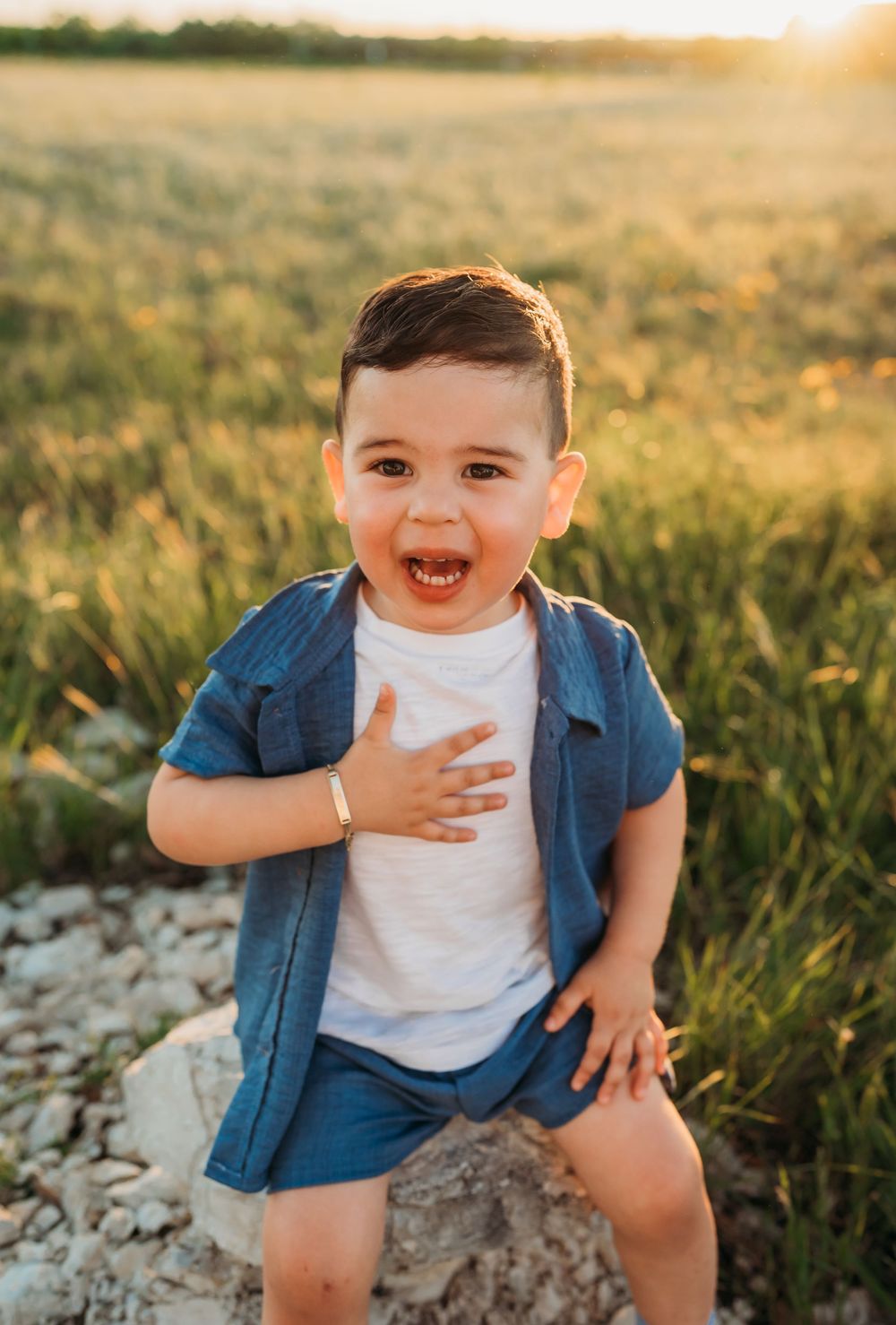 A laughing child wearing a blue button-up shirt and white t-shirt sits on a rock in a sunlit meadow at golden hour.