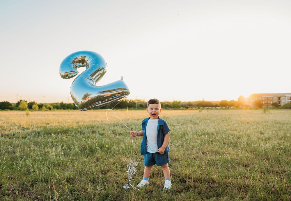 Child stands in grassy field at sunset with blue number 2 balloon floating above in summer celebration.