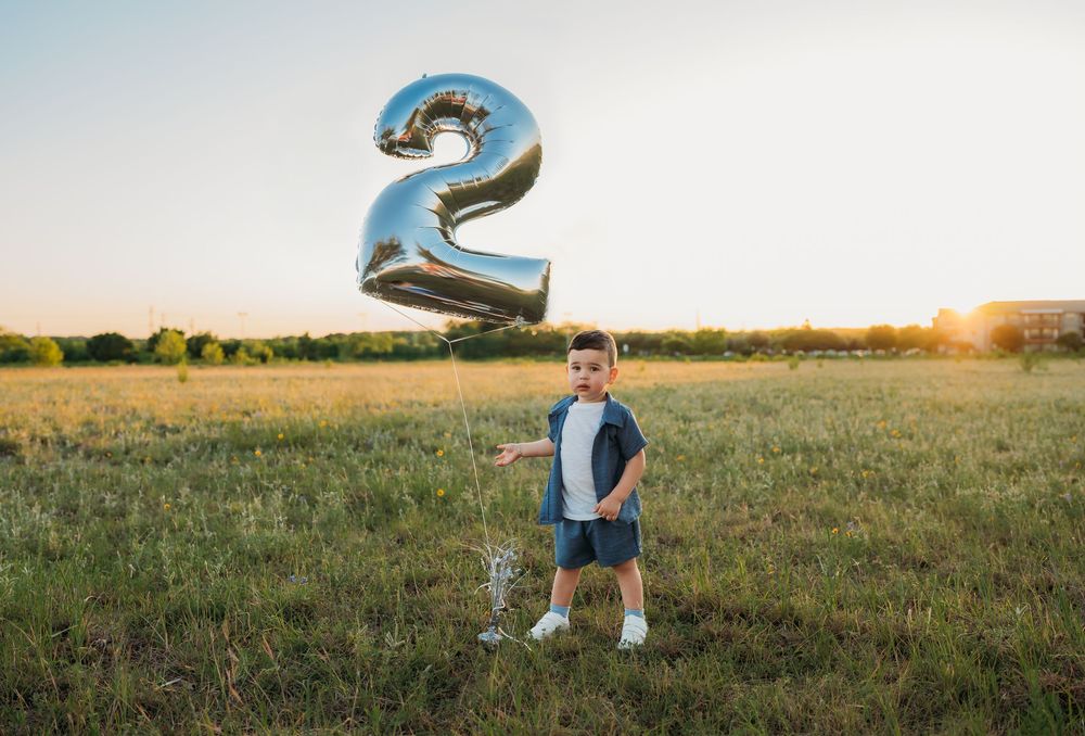 A young child stands in a grassy field at sunset with a large silver number 2 balloon floating above.