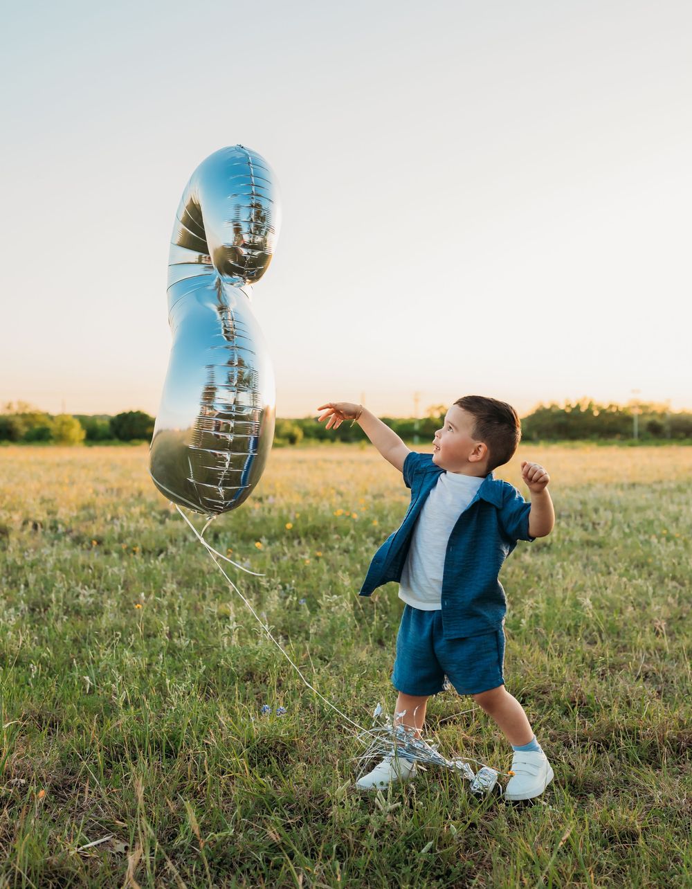 Toddler in blue outfit playing with a large silver balloon in a grassy field at sunset.